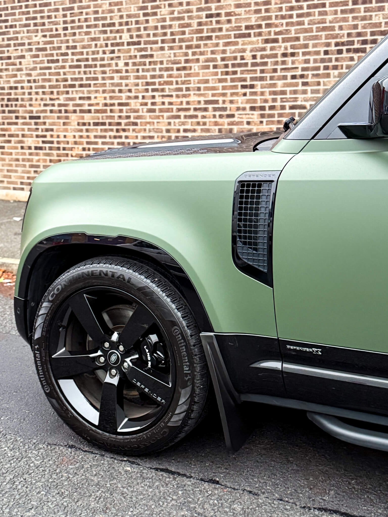A custom Forge Land Rover Defender 90 parked on an asphalt street with a brick wall behind it.