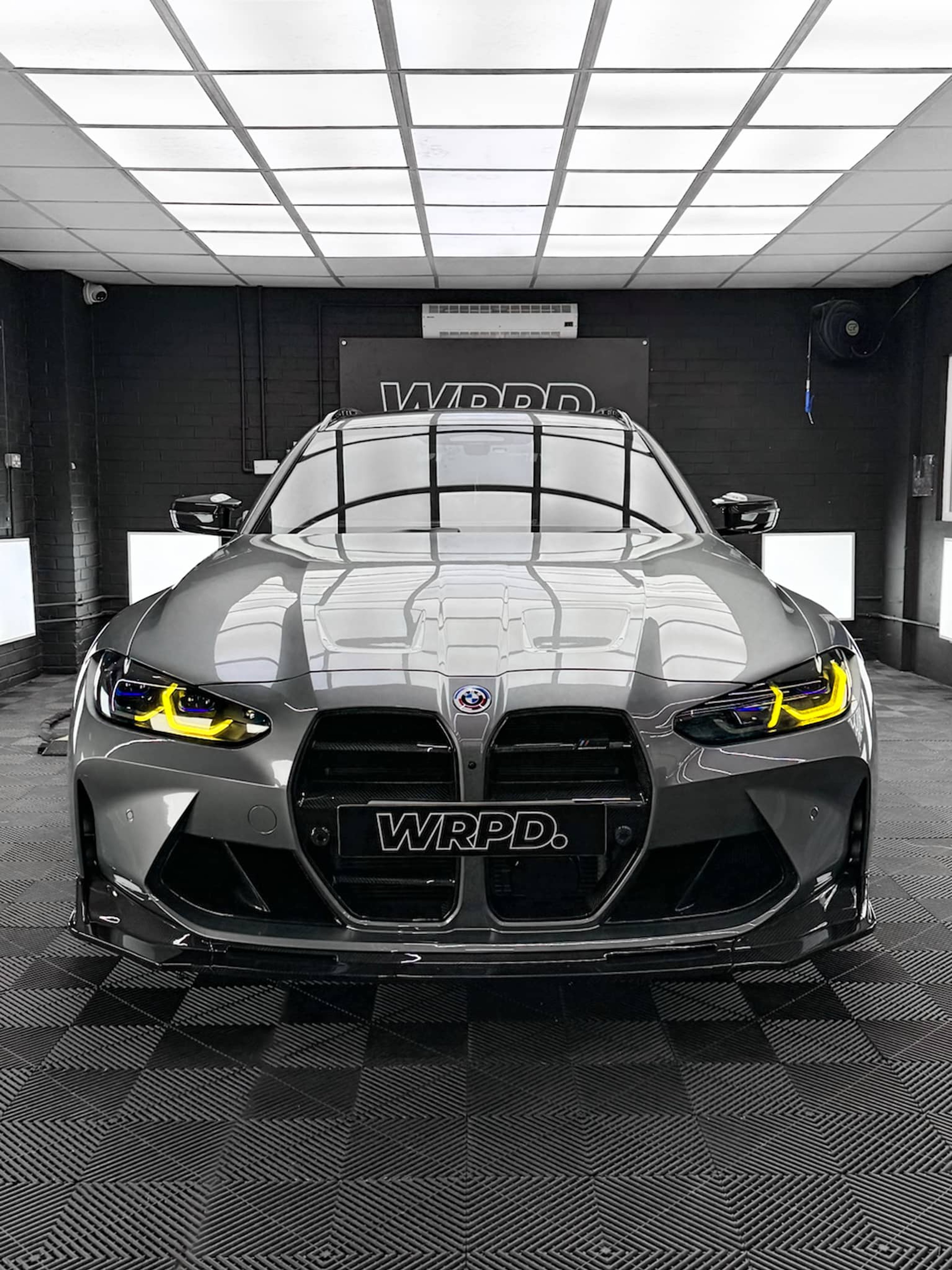 A custom Forge BMW M5 Touring sits centered in a brightly lit, modern black-tiled workshop with overhead square lighting panels.