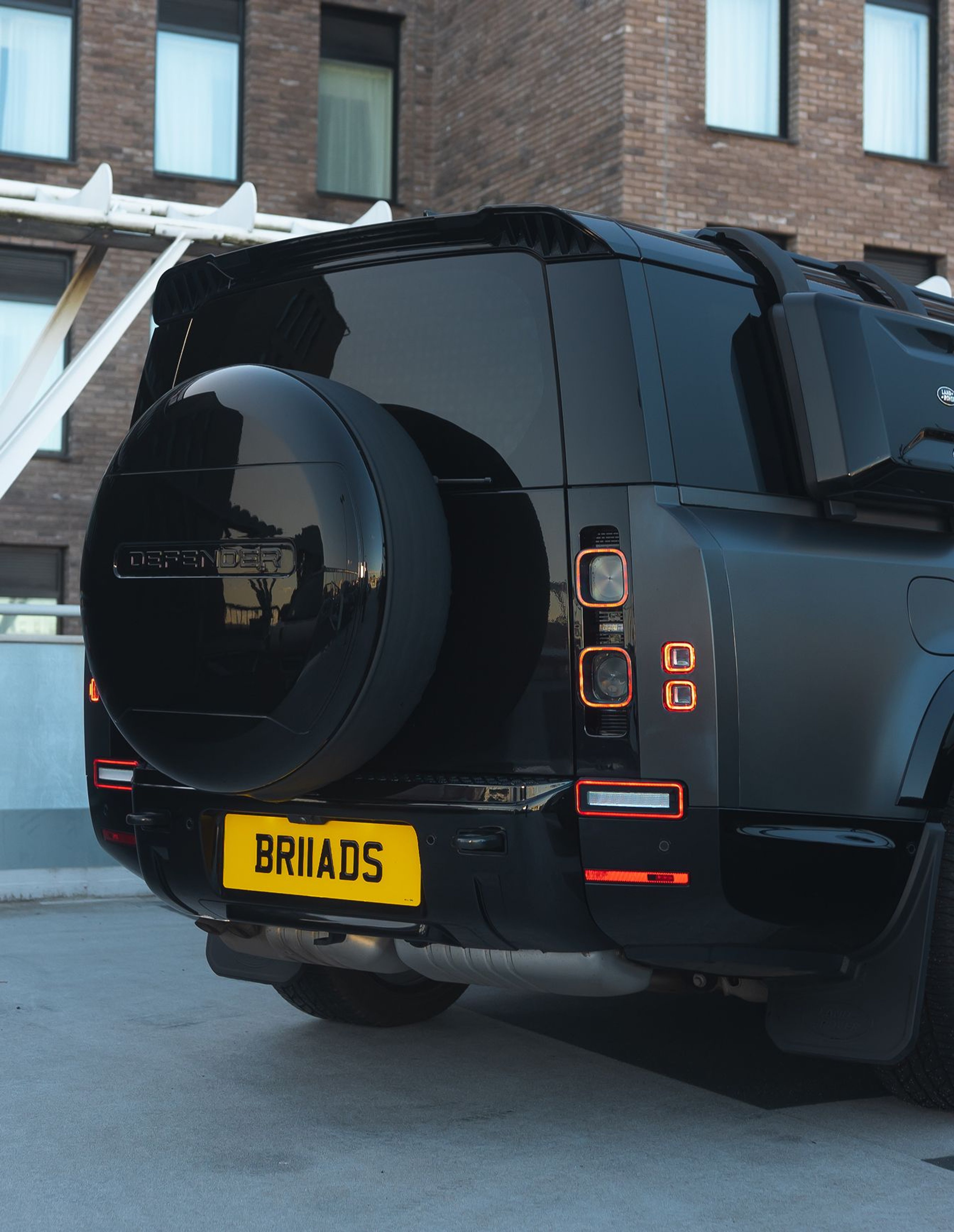 A custom Forge Land Rover Defender 130, in black, is parked with its rear facing a brick building with windows.