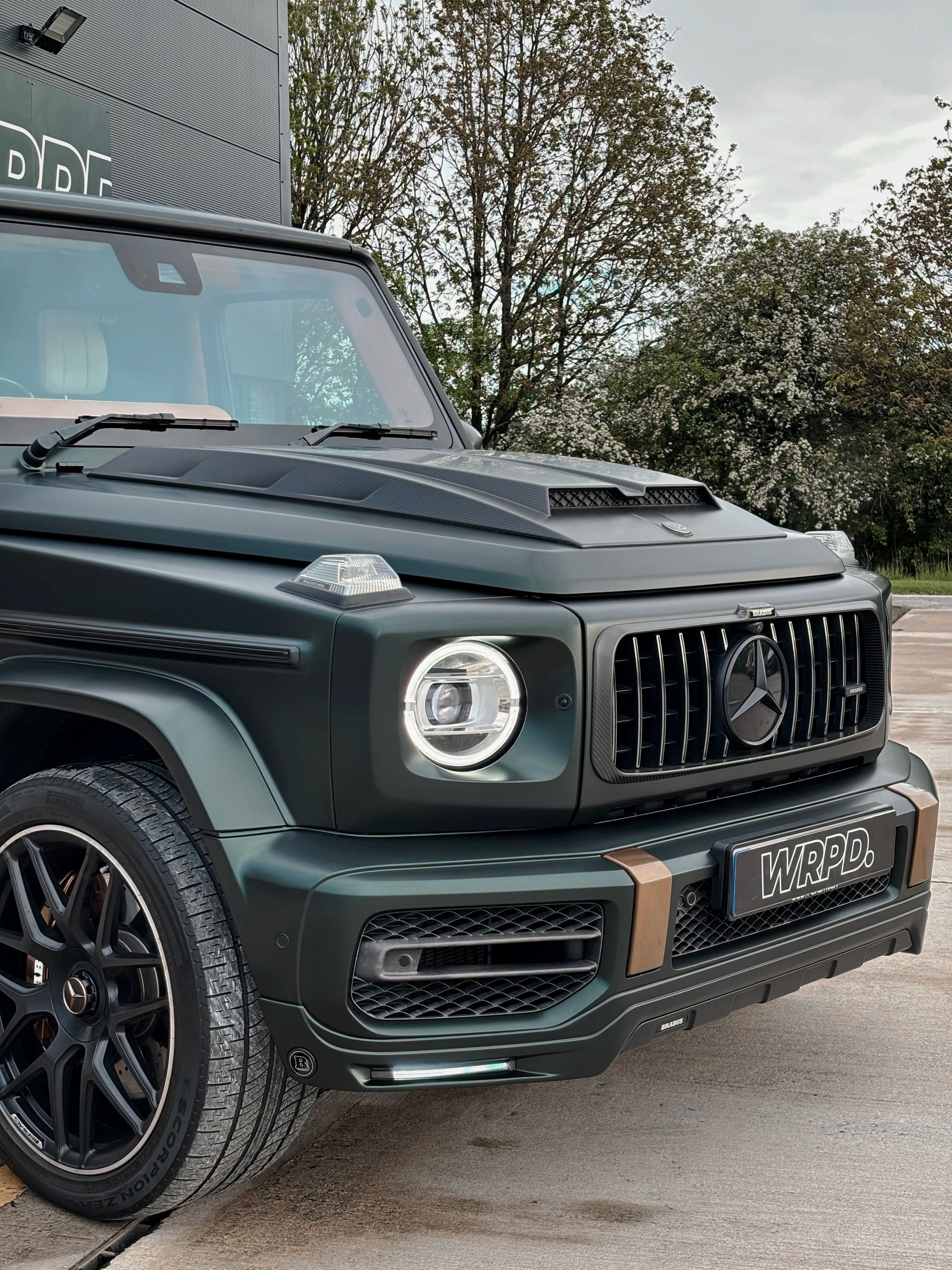 A custom Forge Mercedes G63 with a matte green finish parked on a concrete lot near trees.
