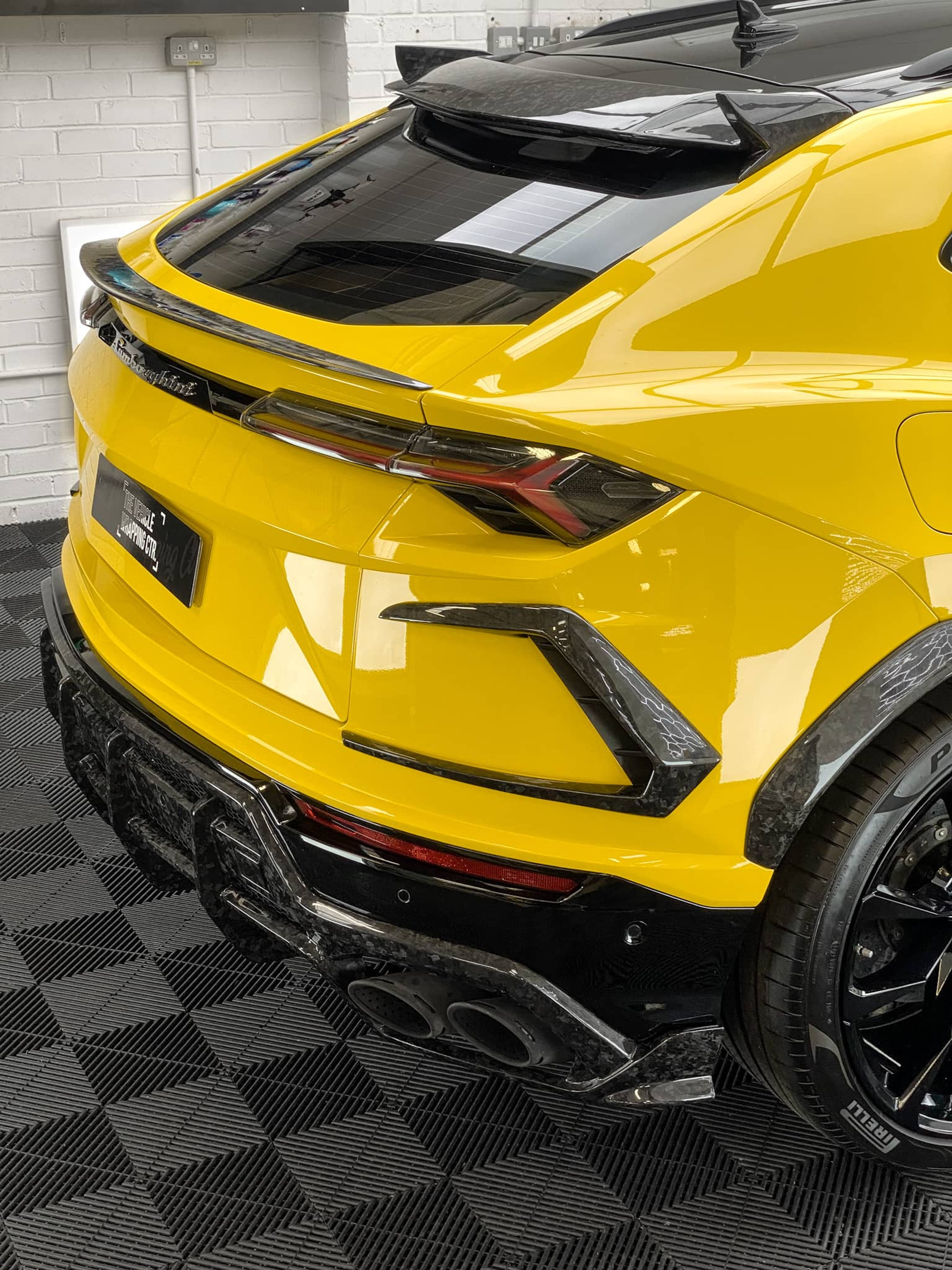 A bright yellow custom Forge Lamborghini Urus parked inside a workshop with patterned black flooring and white brick walls.