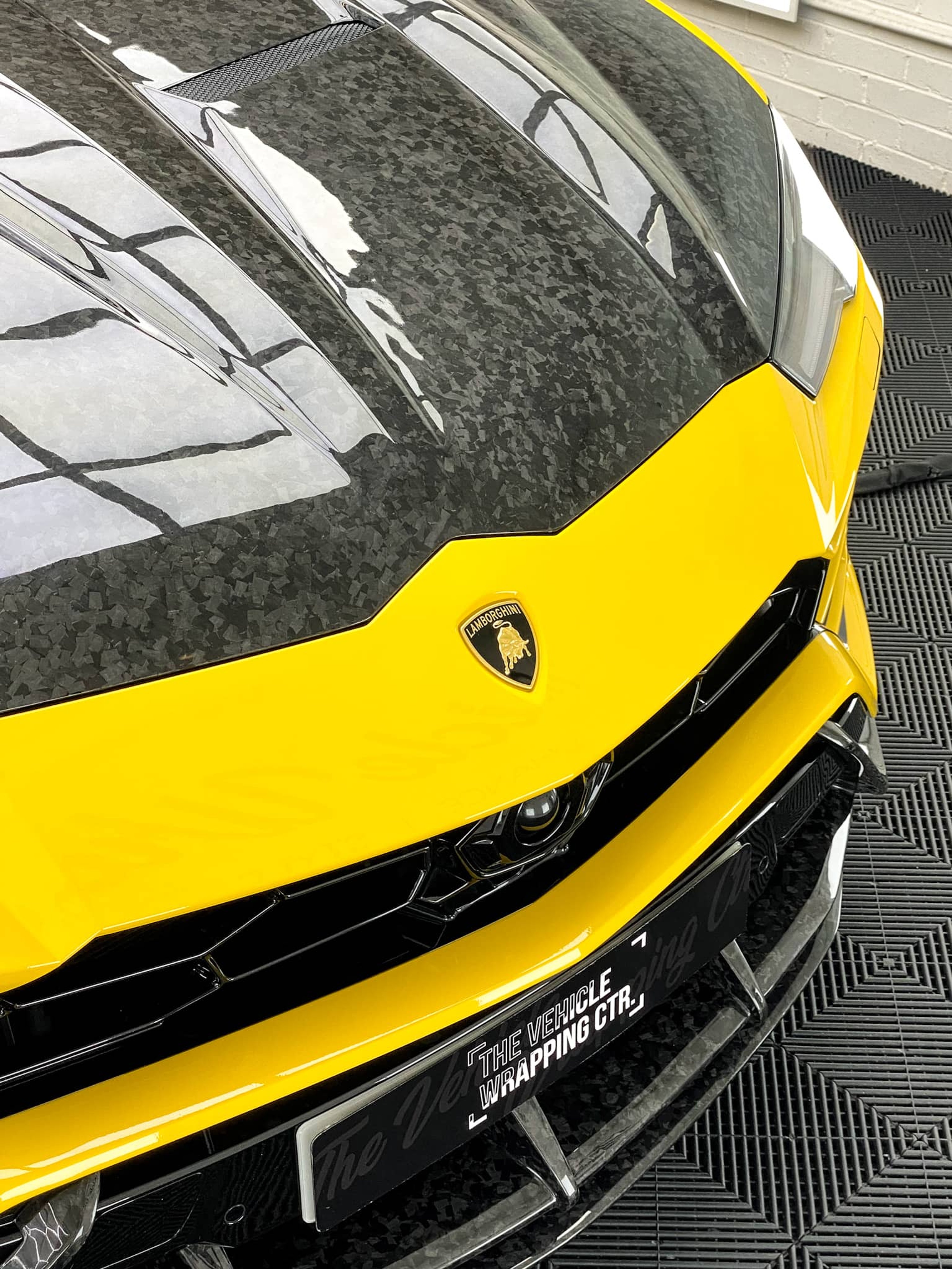 A custom Forge Lamborghini Urus parked on a workshop floor, focusing on its forged carbon hood and yellow front fascia.