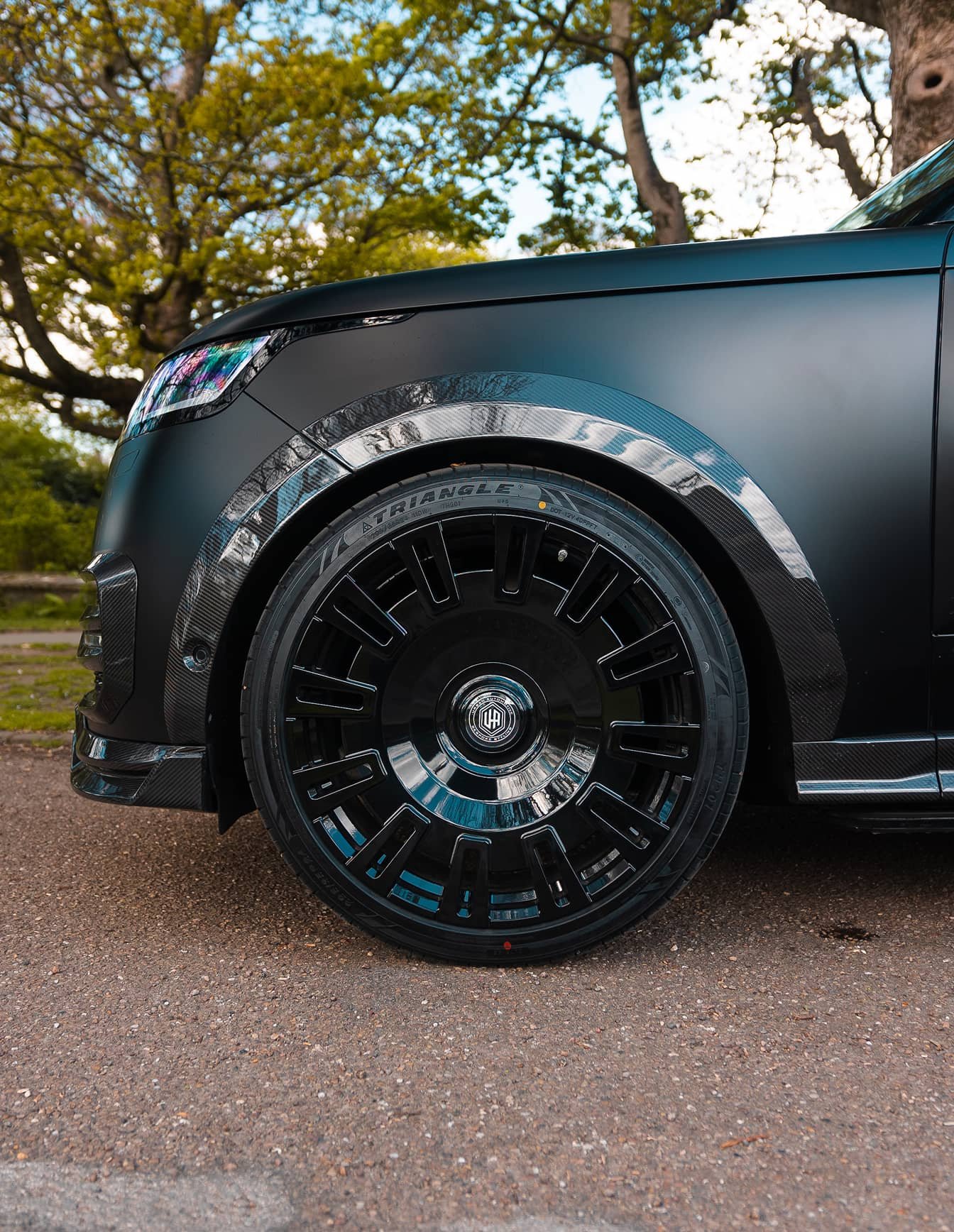 A custom Forge Land Rover Range Rover with matte black paint and carbon fiber accents is parked on an asphalt road under green trees.