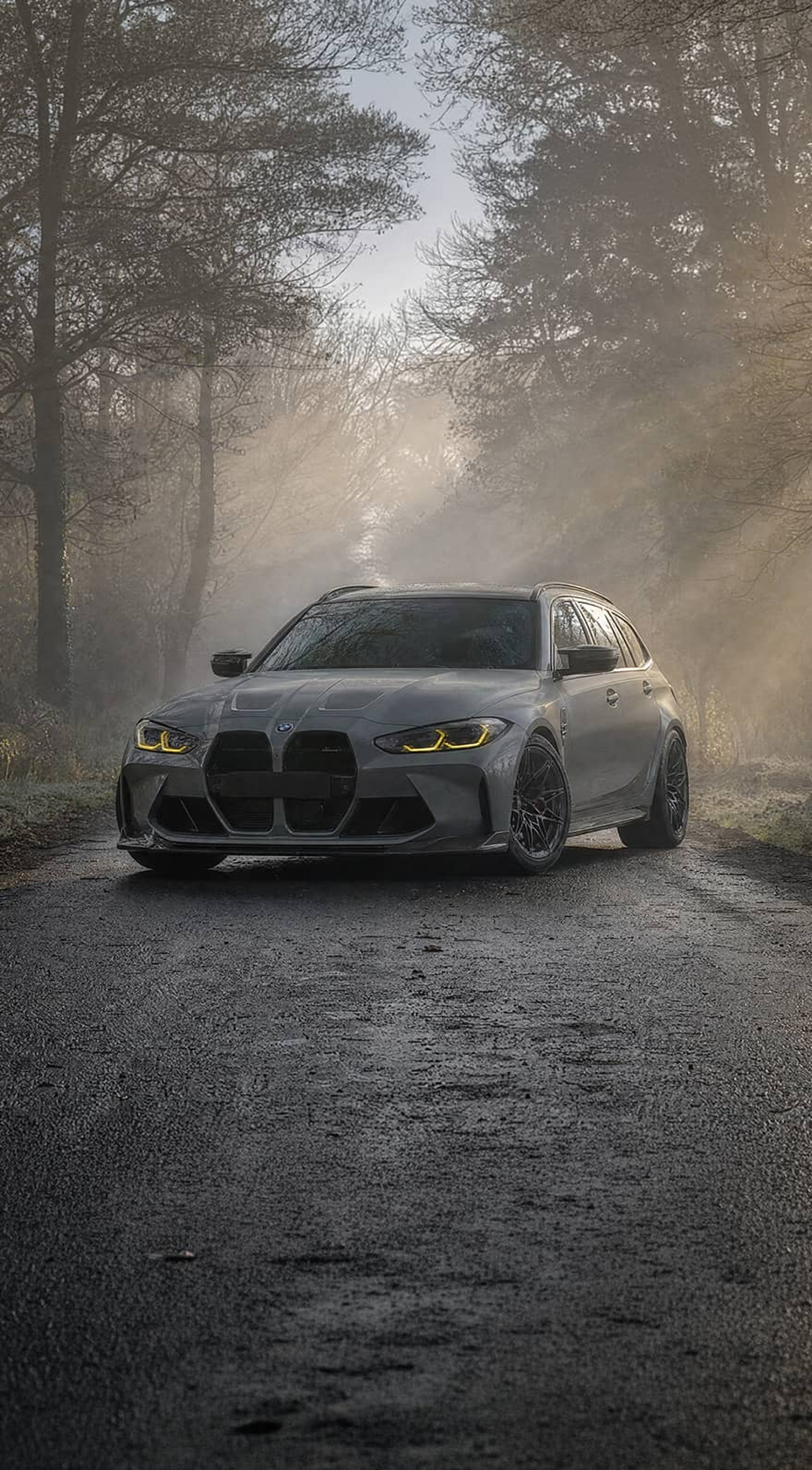 A custom Forge BMW M5 Touring sits on a narrow, wet country road framed by foggy, sunlit trees.