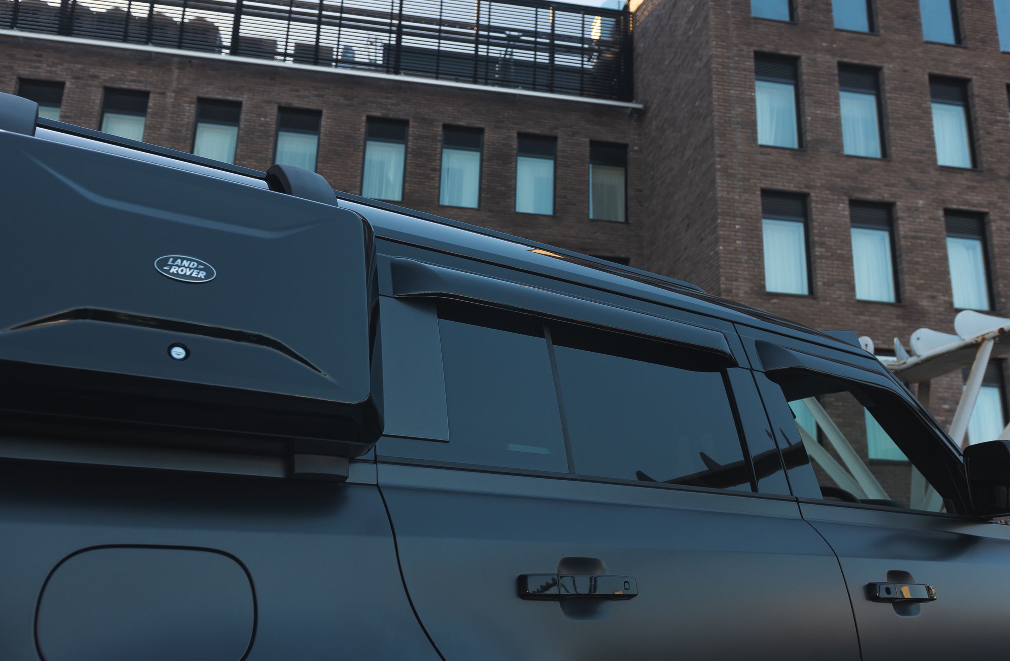 A custom Forge Land Rover Defender 130 with a matte black finish and roof-mounted cargo box is parked in front of a brick building.