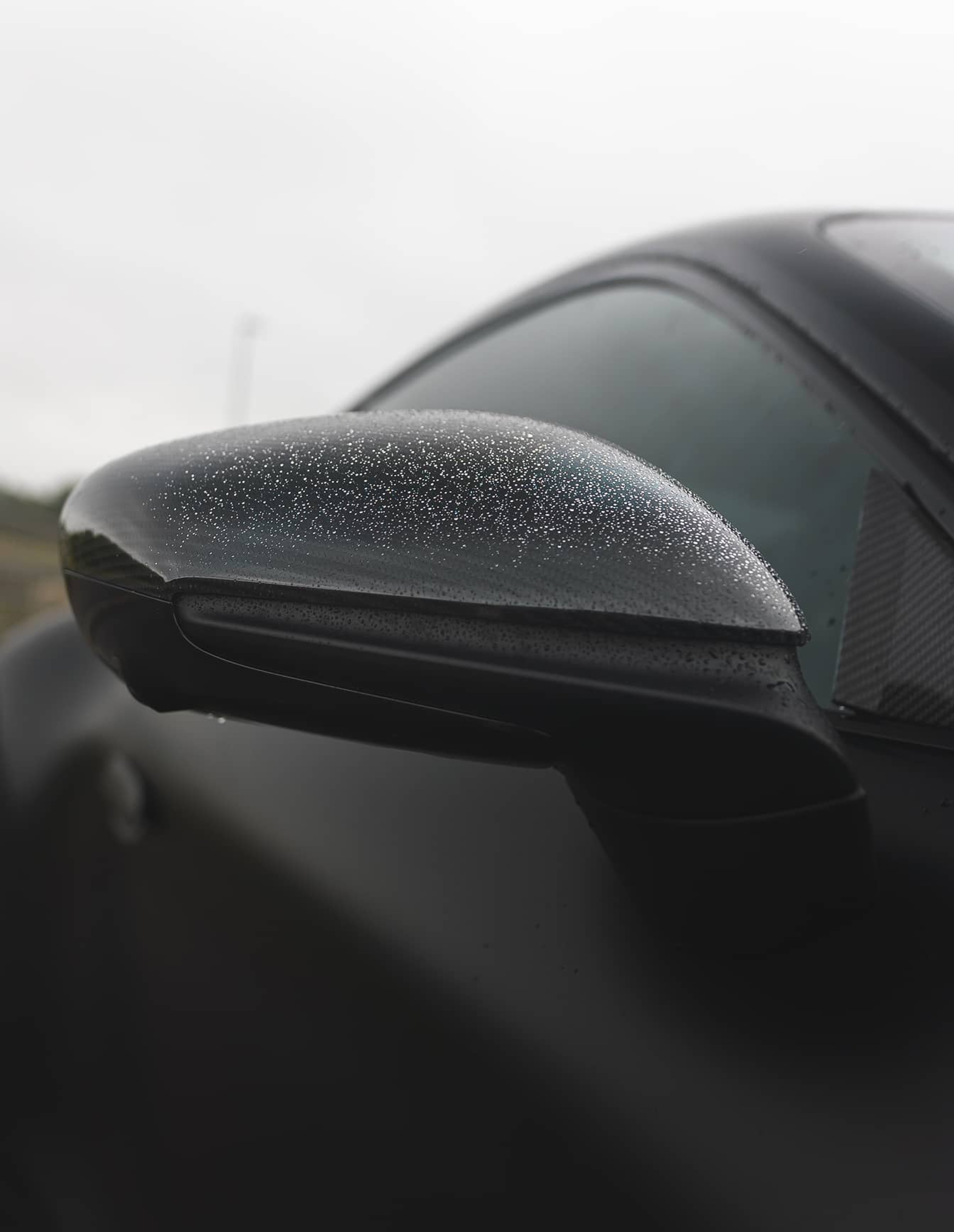 A custom Forge Porsche 911 Carrera Turbo's black side mirror is covered in raindrops on a cloudy day.