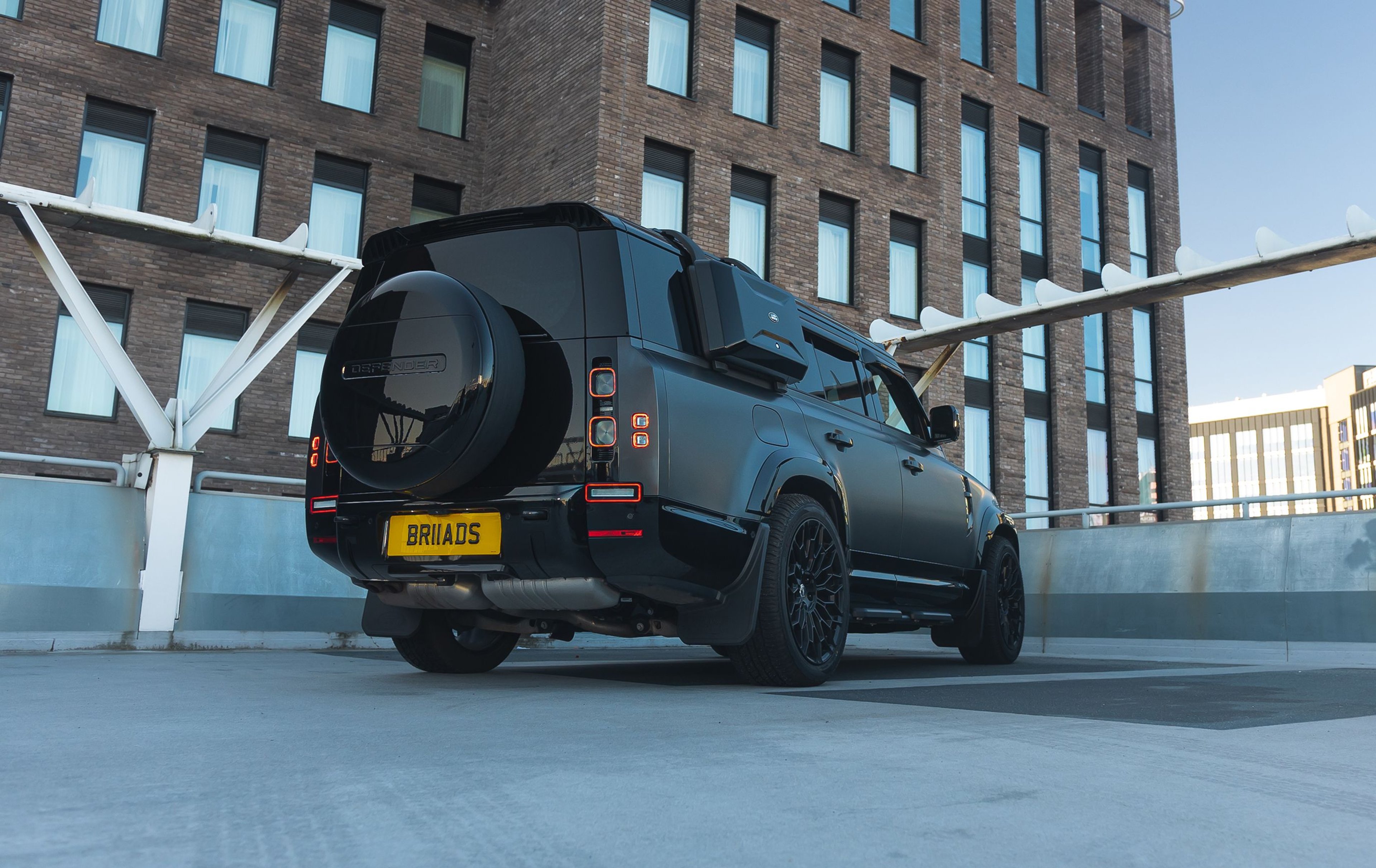 A custom Forge Land Rover Defender 130, matte black, is parked on a concrete rooftop with a brick building in the background.