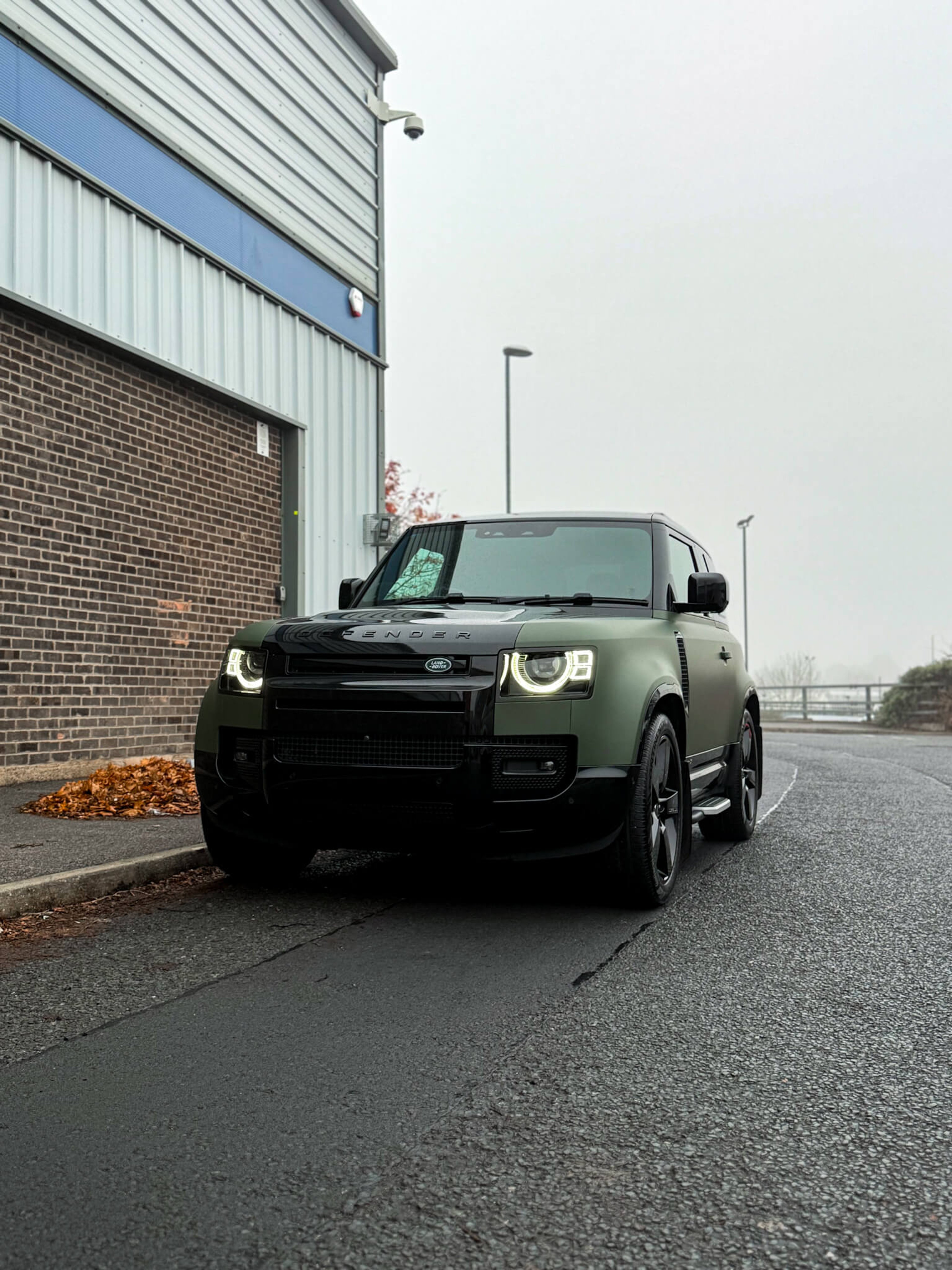 A custom Forge Land Rover Defender 90 parked on an asphalt road beside a brick and metal building on an overcast day.