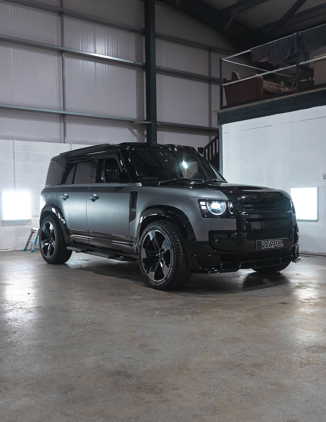 A custom Forge Land Rover Defender 110 with a matte gray and black finish is parked on a concrete floor inside a workshop.