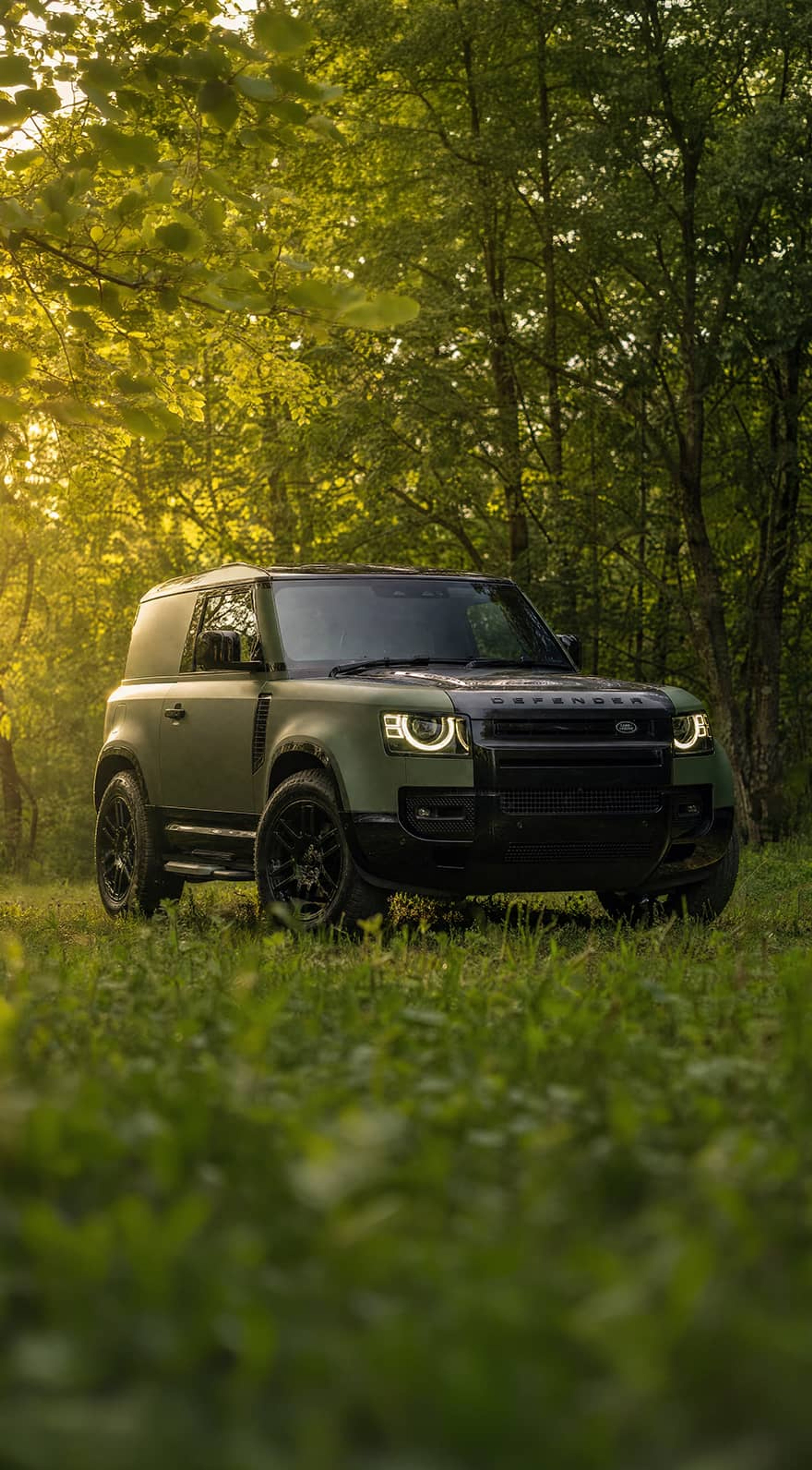 A custom Forge Land Rover Defender 90 parked on a grassy patch in a sunlit forest.