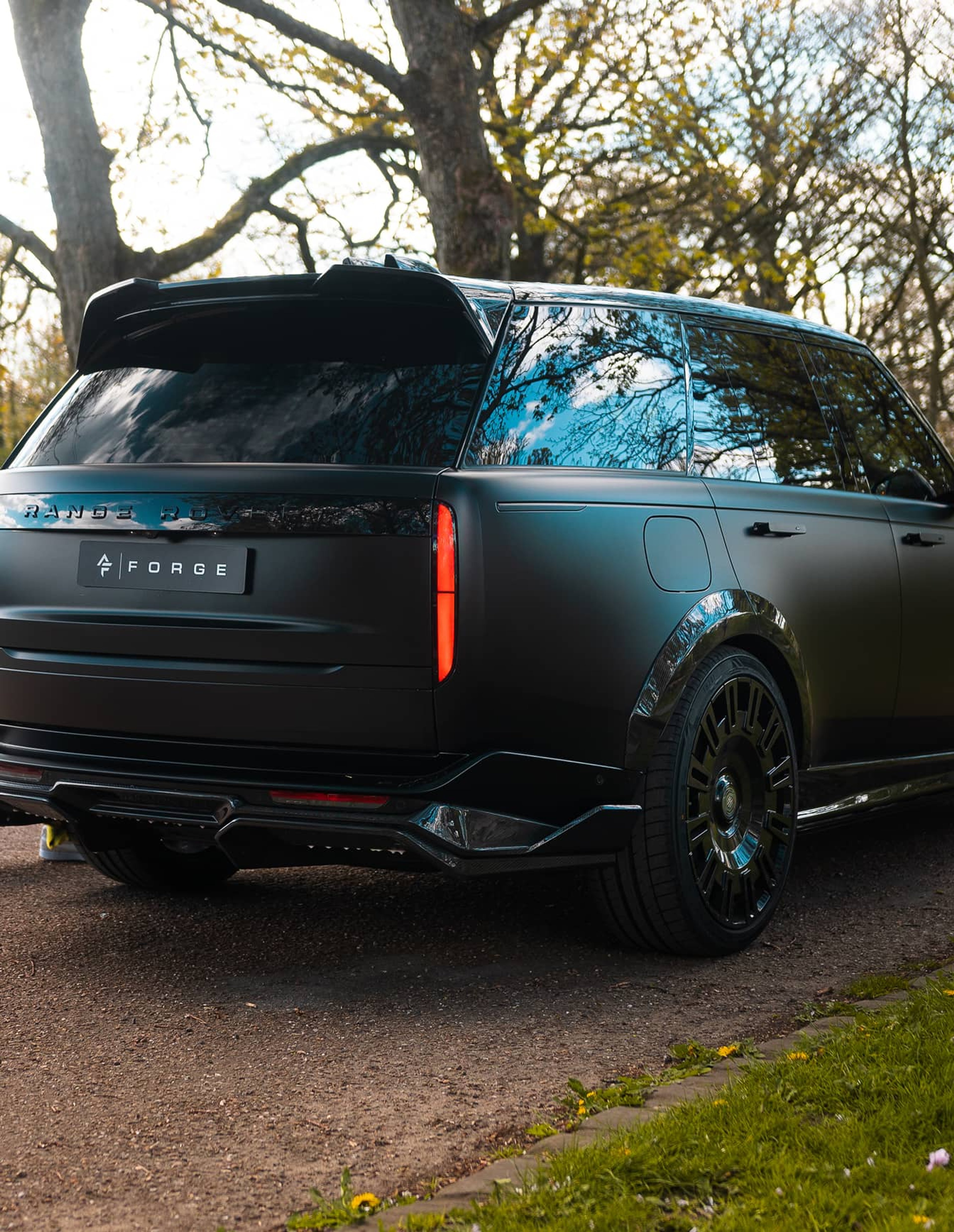 A custom Forge Land Rover Range Rover, matte black, is parked on a gravel path with trees and grass in the background.