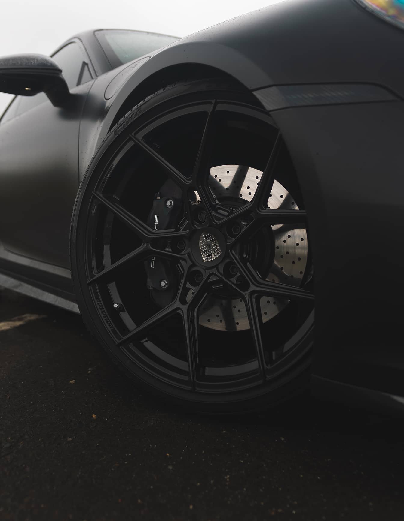 A custom Forge Porsche 911 Carrera Turbo, matte black with glossy black wheels and drilled brake rotors, is parked on a wet surface.