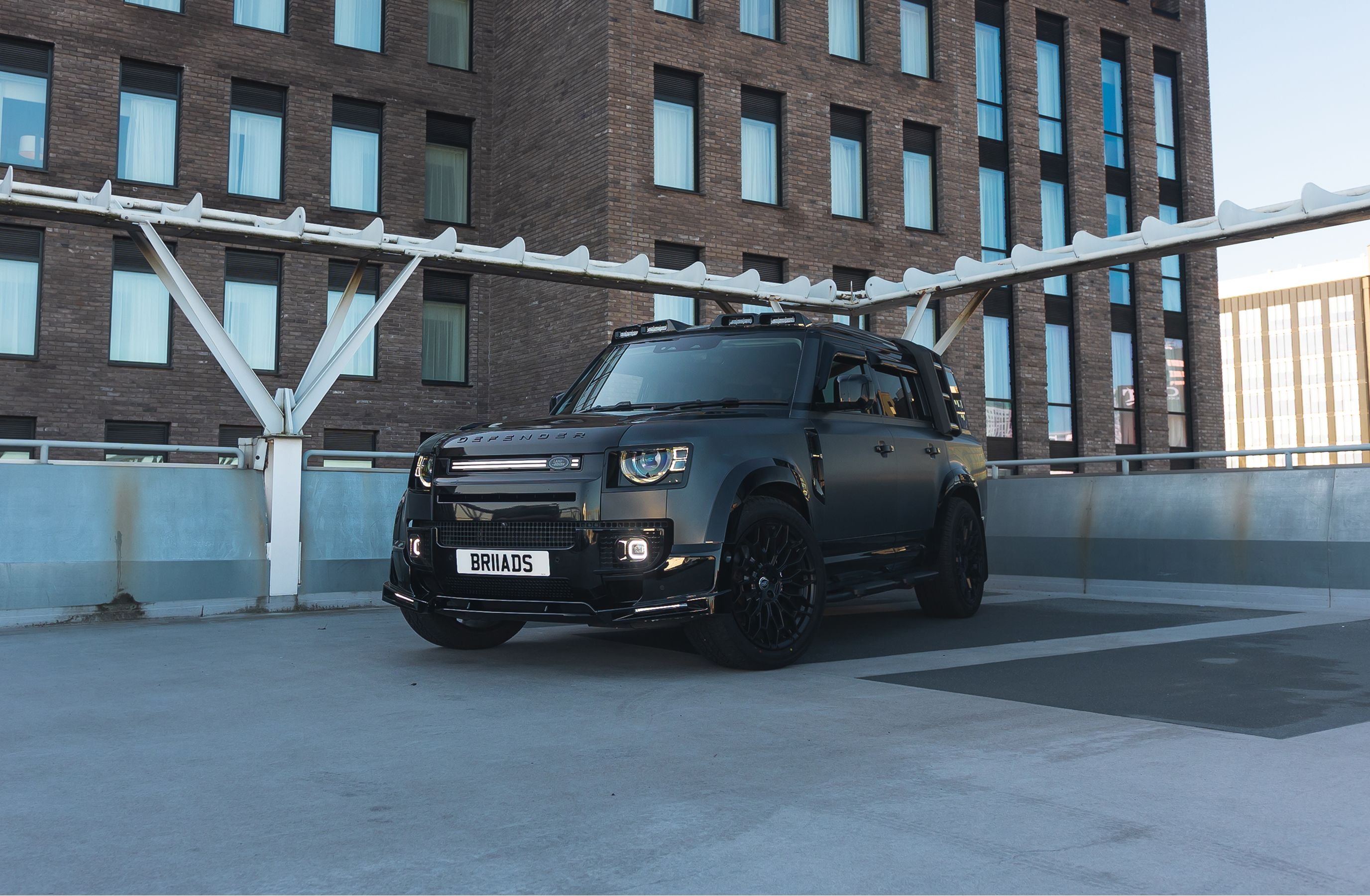 A custom Forge Land Rover Defender 130 in matte black is parked on a concrete rooftop with a brick building in the background.