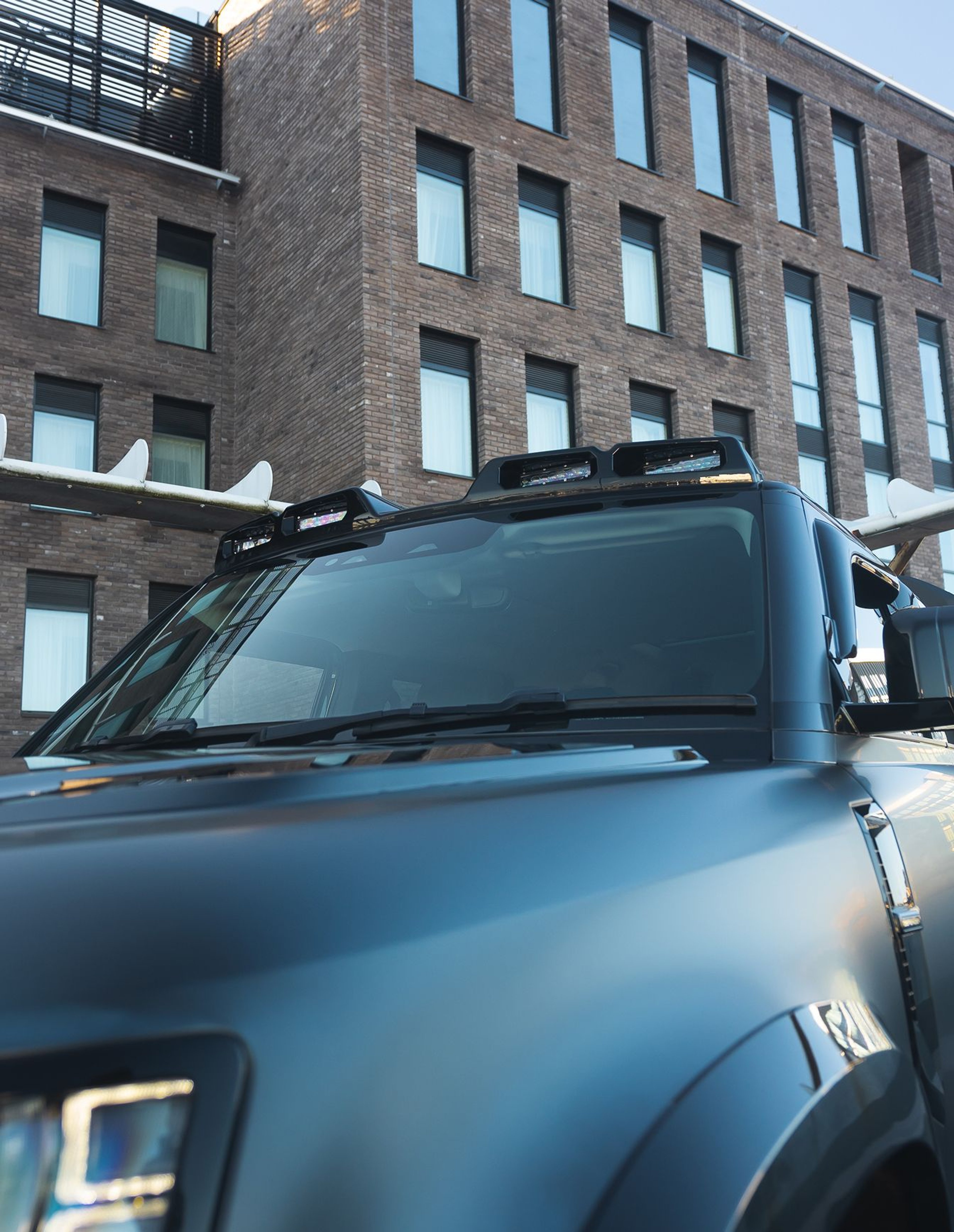 A custom Forge Land Rover Defender 130 with roof-mounted lights and skis, parked in front of a modern brick building.