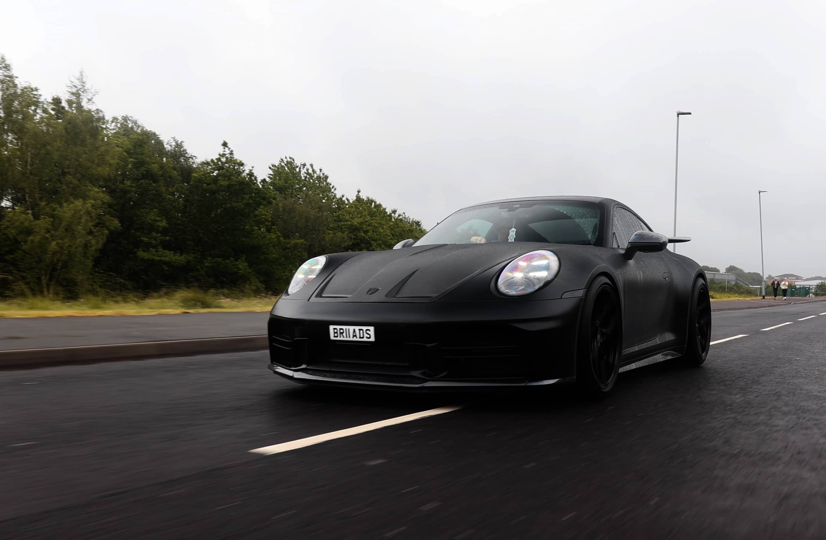 A custom Forge Porsche 911 Carrera Turbo in matte black drives on a wet road under an overcast sky, with trees and streetlights in the background.