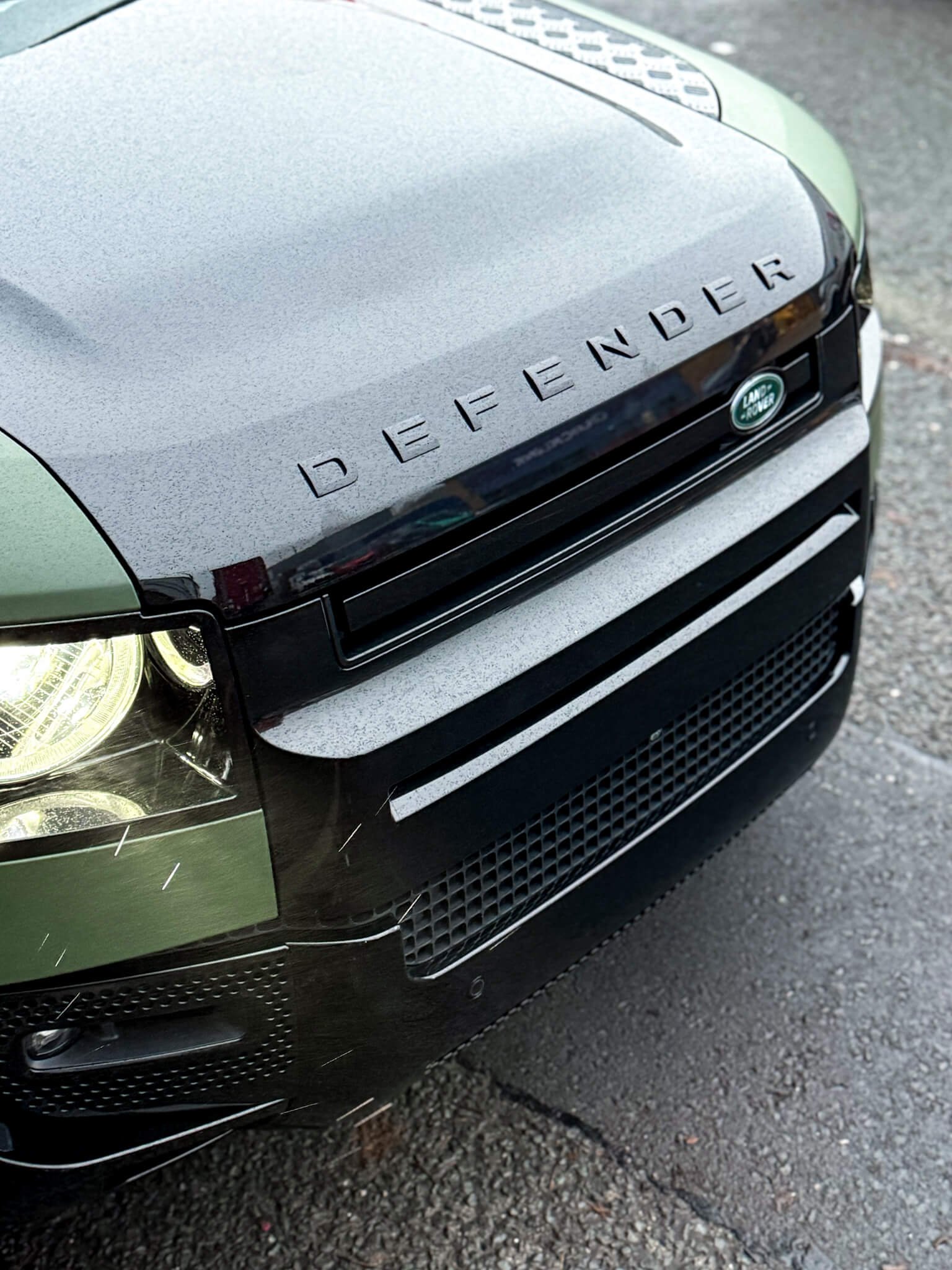 A close-up of the green and black hood on a custom Forge Land Rover Defender 90 parked on a wet asphalt surface.