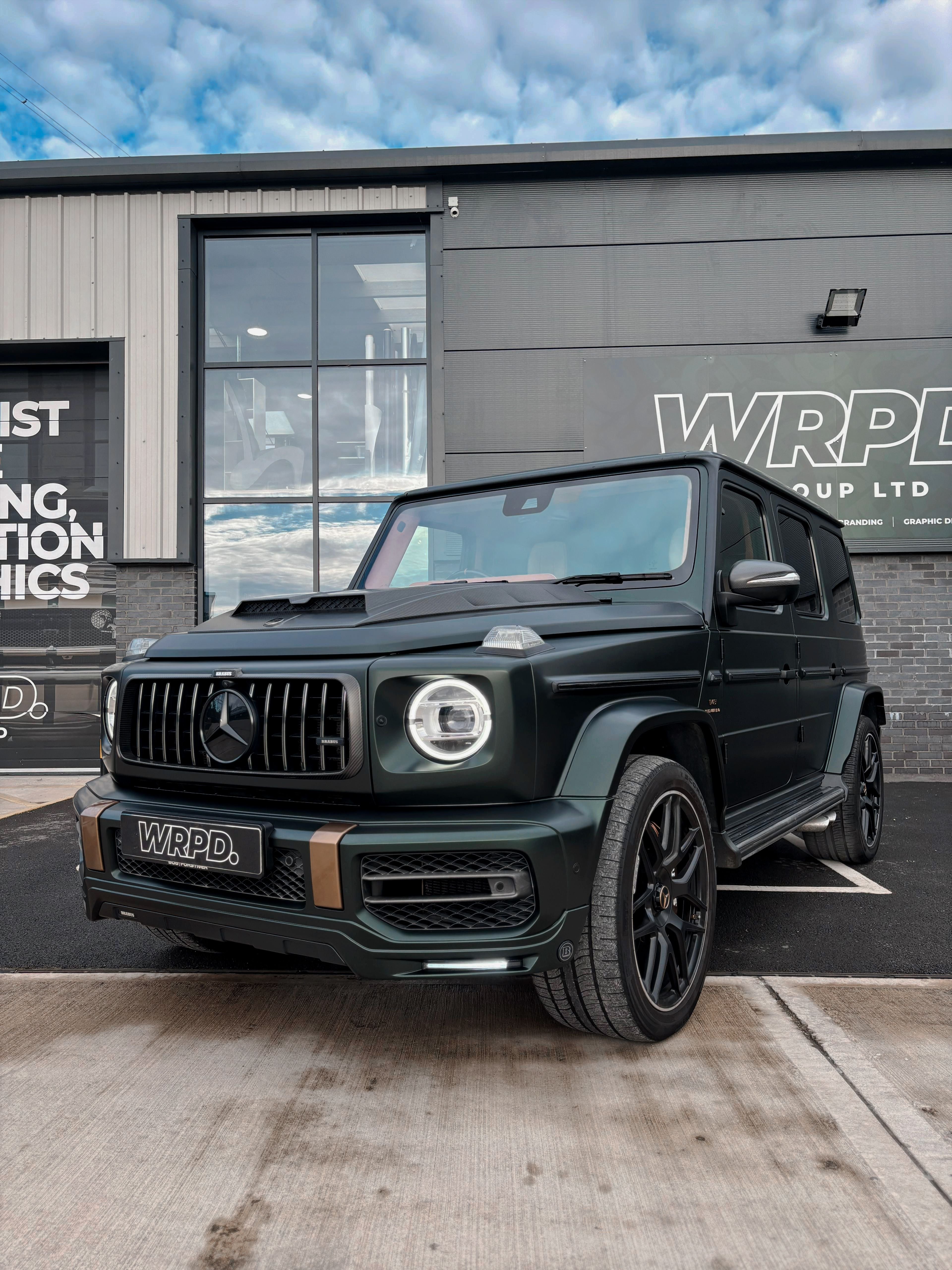 A custom Forge Mercedes G63 in dark green is parked on a wet concrete lot in front of a modern gray building with large windows.